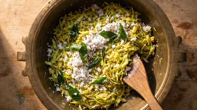 Overhead view of finely shredded cabbage, fresh grated coconut and curry leaves tossed in a bronze uruli pan.