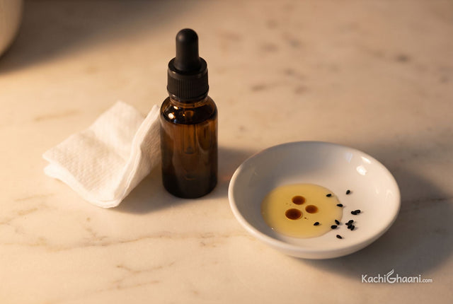 Kalonji oil on skin, a small dark glass dropper bottle of kalonji oil lying beside a folded white cotton pad and a shallow