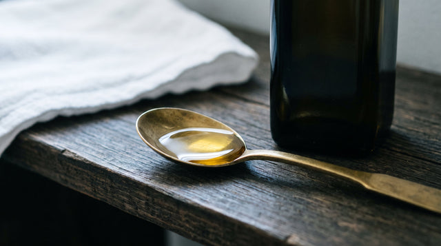 A plain brass spoon filled with golden sesame oil next to an unbranded dark glass bottle on a wooden shelf.