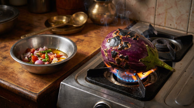 A large whole purple eggplant roasting directly over a blue gas stove flame next to a bowl of chopped onions and tomatoes.